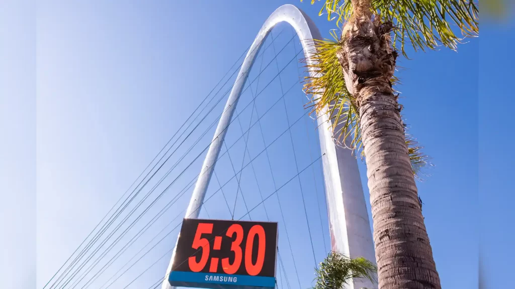 Blue clear sky, with a palm tree in the front. In the background there is a clock stating that it's 5:30 and a giant metal arch with cables.