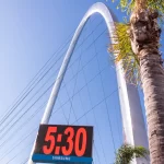 Blue clear sky, with a palm tree in the front. In the background there is a clock stating that it's 5:30 and a giant metal arch with cables.