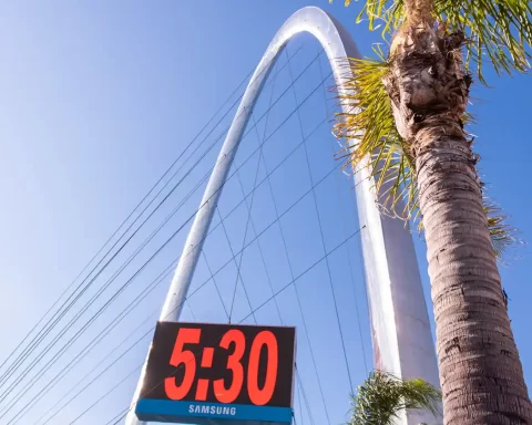 Blue clear sky, with a palm tree in the front. In the background there is a clock stating that it's 5:30 and a giant metal arch with cables.