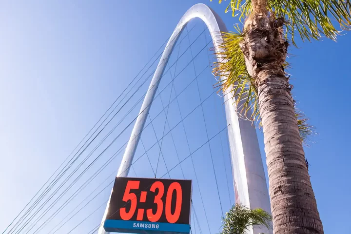 Blue clear sky, with a palm tree in the front. In the background there is a clock stating that it's 5:30 and a giant metal arch with cables.